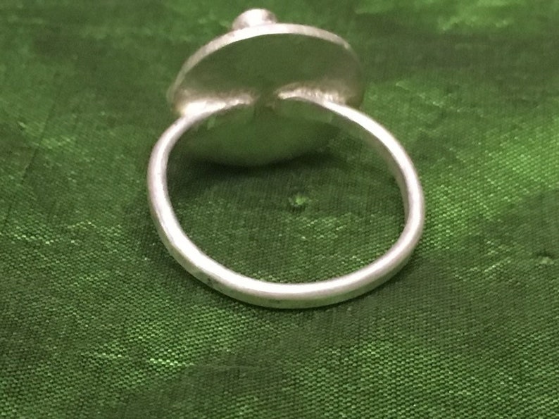 May include: A silver ring with a round, flat top. The ring is on a green fabric background.