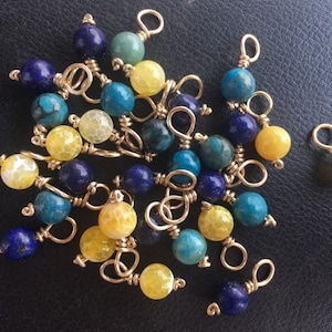 Könnte beinhalten: Eine Sammlung von Edelsteinperlen mit goldenen Drahtschlaufen. Die Perlen sind gelb, blau und türkis.