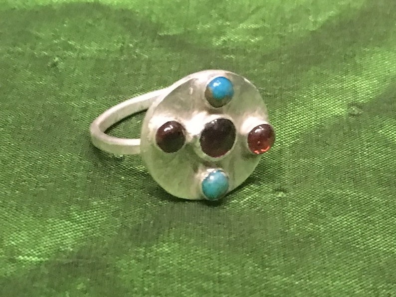 May include: A silver ring with a round face set with five stones. Three of the stones are dark red and two are turquoise.