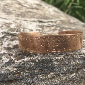 Könnte beinhalten: Ein Kupferarmband mit einem geometrischen Muster aus Dreiecken und Kreisen. Das Armband liegt auf einer Holzoberfläche.