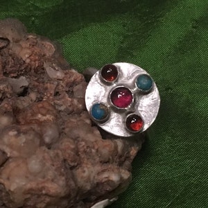 May include: A silver ring with a round, textured setting featuring five colorful gemstones. The ring has a red, pink, and two orange gemstones, and a turquoise gemstone.