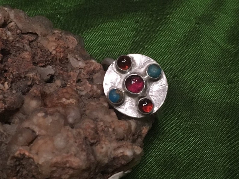 May include: A silver ring with a round, textured setting featuring five colorful gemstones. The ring has a red, pink, and two orange gemstones, and a turquoise gemstone.