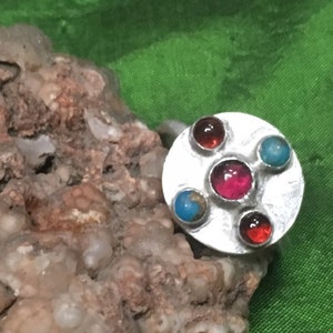 May include: A silver ring with a round, textured setting featuring four gemstones: two red, one pink, and one turquoise.