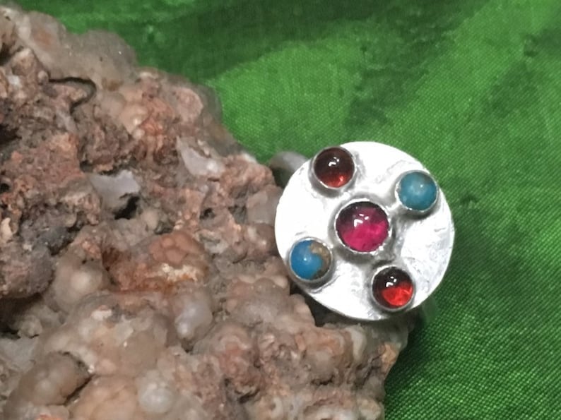 May include: A silver ring with a round, textured setting featuring four gemstones: two red, one pink, and one turquoise.