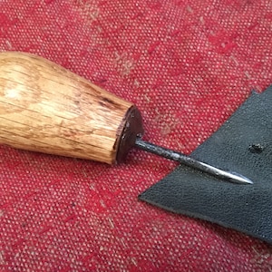 May include: A leather stitching awl with a wooden handle and a sharp metal point. The awl is resting on a piece of dark leather.