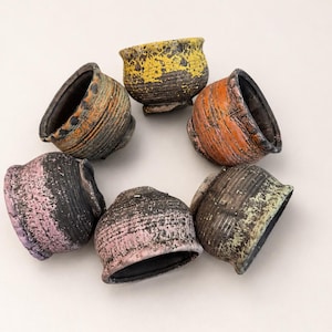May include: Six small, round ceramic bowls arranged in a circle. Each bowl features a dark, textured base with horizontal ridges and a unique, distressed-look glaze in yellow, orange, pink, green, and multi-colored earth tones on the upper rim.