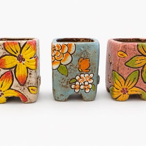May include: Three square ceramic planters with rounded corners and four small feet. The left planter is cream with yellow flowers, the center is light blue with orange and white floral patterns, and the right is pink with yellow flowers.