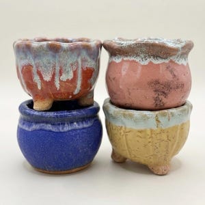 May include: Four small ceramic pots with three feet each. The top left pot is orange with a blue drip glaze. The top right pot is pink with a white rim. The bottom left pot is solid blue. The bottom right pot is yellow with a white rim.