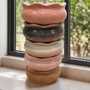 May include: A vertical stack of five ceramic bowls with scalloped edges. The bowls feature a glossy finish in various colors including light pink, dark charcoal, cream, soft pink, and a warm tan, each with subtle tonal variations in the glaze.