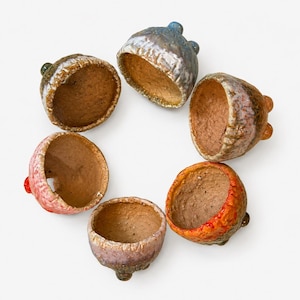 May include: Six small, bowl-shaped ceramic vessels arranged in a circle. Each vessel features a textured, glazed exterior in varying colors including blue, pink, orange, and brown, with a rough, unglazed, light brown interior surface.