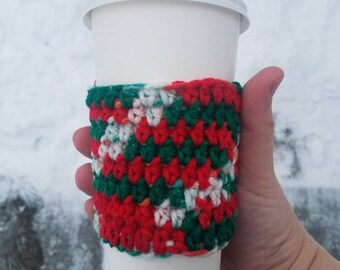 May include: A white to-go coffee cup with a red, green, and white crocheted cozy. The cozy has a geometric pattern.
