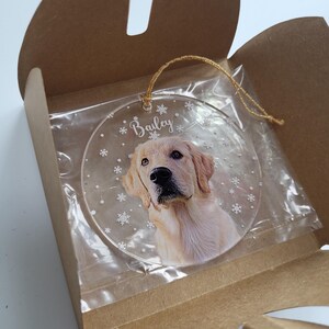 Clear round Christmas ornament with a dog photo and name with snow flakes background placed in the middle an opened brown gift envelop