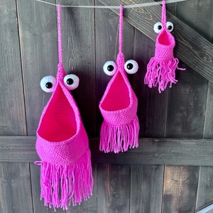 May include: Three pink crocheted hanging baskets with white googly eyes and pink fringe. The baskets are hanging from a white string against a dark wood background.