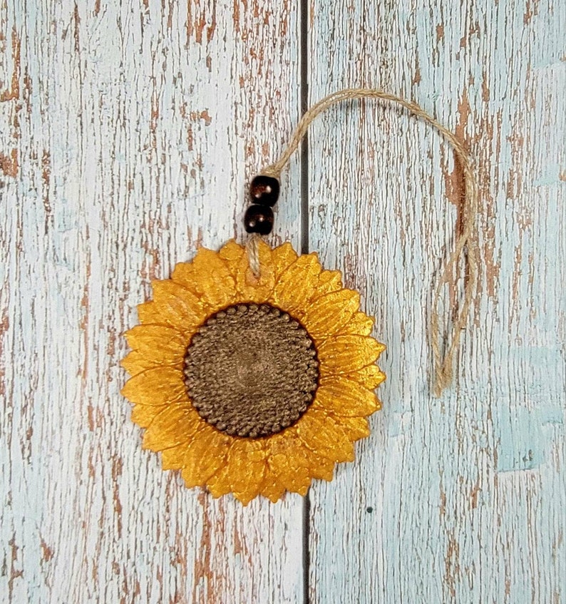 Puede incluir: Un girasol de color dorado con un centro marr&oacute;n, colgado de una cuerda de yute con cuentas marrones. El girasol est&aacute; sobre un fondo de madera blanqueada.
