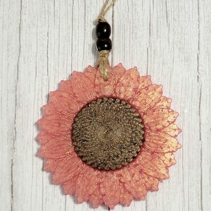 Puede incluir: Un adorno de girasol de purpurina rosa y dorada con un colgador de cuerda marr&oacute;n. El girasol tiene un centro texturizado y es un acento decorativo para un hogar.