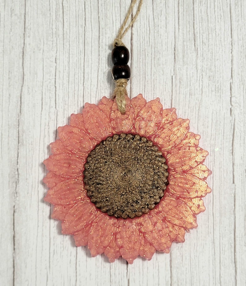 Puede incluir: Un adorno de girasol de purpurina rosa y dorada con un colgador de cuerda marr&oacute;n. El girasol tiene un centro texturizado y es un acento decorativo para un hogar.