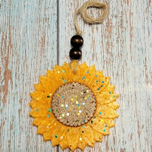 Puede incluir: Un ambientador de coche en forma de girasol amarillo con detalles de purpurina y una cuerda marr&oacute;n para colgar.