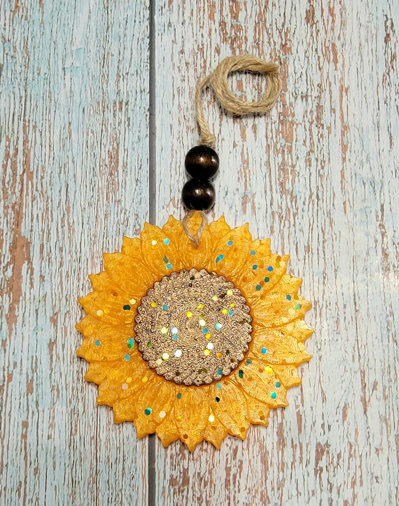 Puede incluir: Un ambientador de coche en forma de girasol amarillo con detalles de purpurina y una cuerda marr&oacute;n para colgar.
