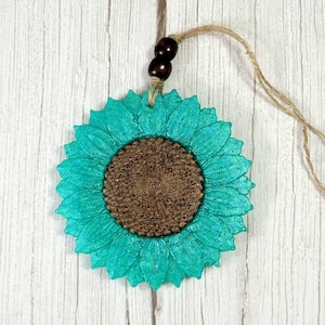 Puede incluir: Un adorno de girasol azul turquesa con un centro marr&oacute;n. El adorno cuelga de una cuerda de yute con cuentas de madera marr&oacute;n.