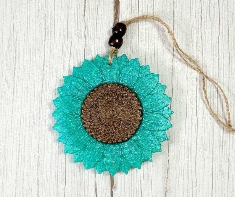 Puede incluir: Un adorno de girasol azul turquesa con un centro marr&oacute;n. El adorno cuelga de una cuerda de yute con cuentas de madera marr&oacute;n.