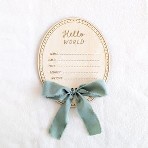 Può includere: Cartello ovale in legno per l'annuncio del bambino con le parole "Hello WORLD" incise in alto. Il cartello ha un bordo a puntini e linee per nome, data, ora, lunghezza e peso. Un nastro verde salvia è legato in basso.