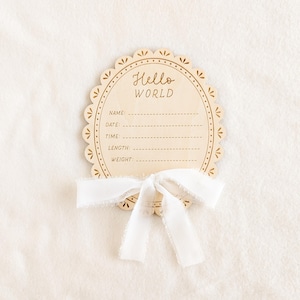 May include: A wooden baby announcement plaque with the words "Hello World" in a decorative font. The plaque has space to write the baby's name, date, time, length, and weight. A white ribbon is tied at the bottom.