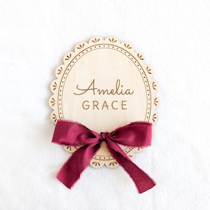 Könnte beinhalten: Ein Holzschild mit gewelltem Rand, graviert mit dem Namen "Amelia Grace". Unten ist eine bordeauxfarbene Schleife gebunden. Die Plakette ist hellbraun und das Band hat einen ausgefransten Rand.