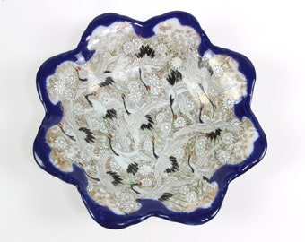 May include: A white ceramic bowl with a blue rim and a design of white cranes flying over a field of gold flowers. The bowl has a scalloped edge.