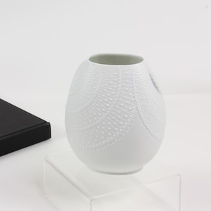 May include: A white ceramic vase with a rounded, egg-like shape. The exterior surface features a raised, textured pattern of radiating lines and small dots that create a fan-like design across the front and side of the vessel.