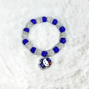 May include: A blue and silver beaded bracelet with a charm. The bracelet has alternating blue and silver beads. A charm of a cartoon character with a red bow hangs from the bracelet. The bracelet is displayed on a white, fluffy surface.