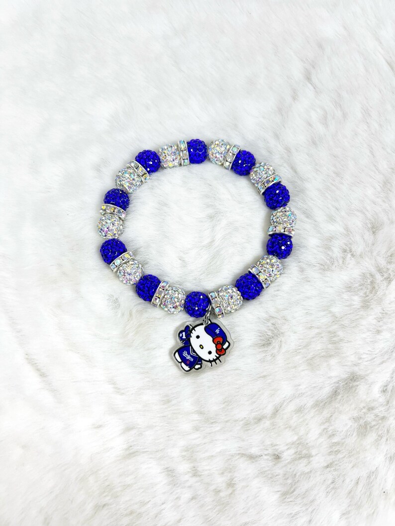 May include: A blue and silver beaded bracelet with a charm. The bracelet has alternating blue and silver beads. A charm of a cartoon character with a red bow hangs from the bracelet. The bracelet is displayed on a white, fluffy surface.