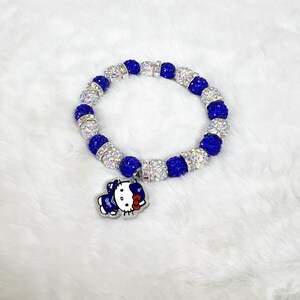 May include: A bracelet with alternating blue and clear crystal beads. A charm of a cartoon character in blue and white is attached. The bracelet is displayed on a white, fluffy surface.