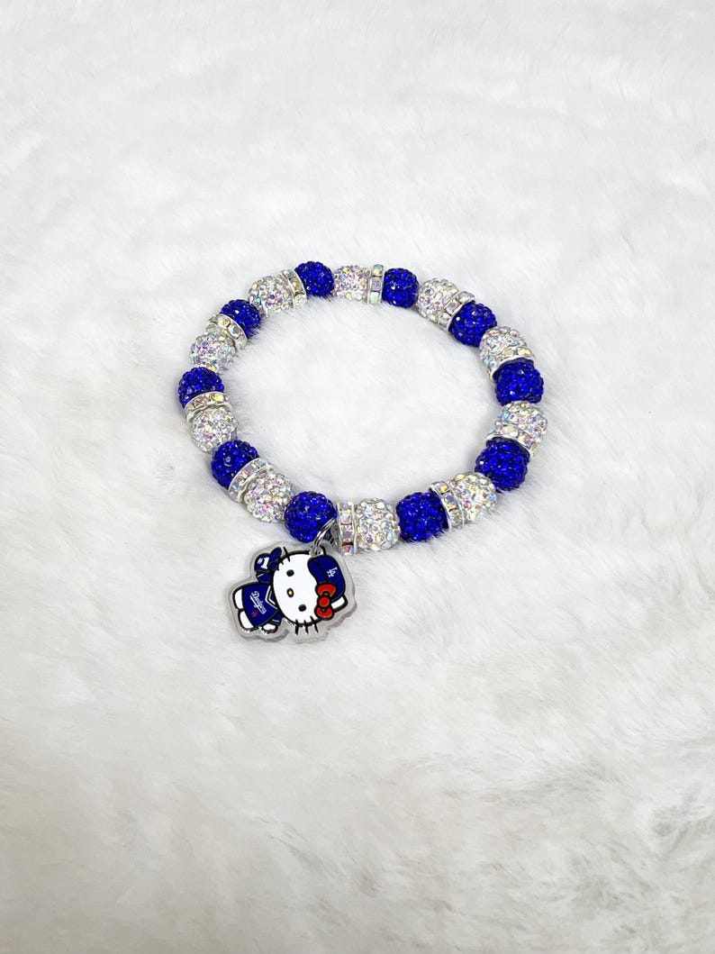 May include: A bracelet with alternating blue and clear crystal beads. A charm of a cartoon character in blue and white is attached. The bracelet is displayed on a white, fluffy surface.