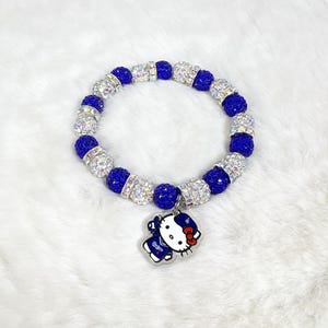 May include: A bracelet with alternating blue and silver beads. A charm of a cartoon character in a blue uniform with white details and a red bow is suspended from the bracelet.