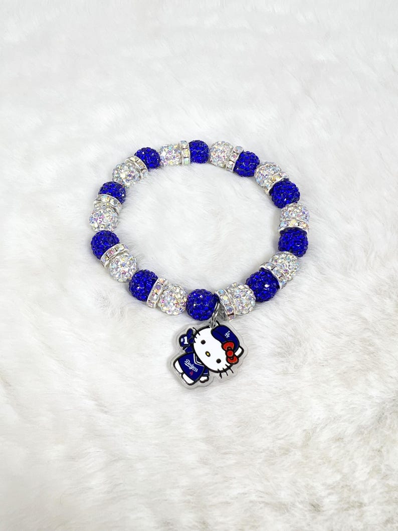 May include: A bracelet with alternating blue and silver beads. A charm of a cartoon character in a blue uniform with white details and a red bow is suspended from the bracelet.
