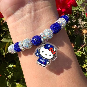 May include: A bracelet with blue and silver beads, and a charm of Hello Kitty in a Dodgers baseball uniform. The bracelet features a mix of round and faceted beads.