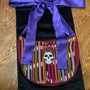 Puede incluir: Una bolsa de tela a rayas de colores con un bordado de calavera y flores. La bolsa tiene un asa de cinta de satén morado.