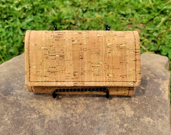 May include: A small, rectangular cork wallet with a gold flecked pattern. The wallet is closed and sitting on a brown surface.
