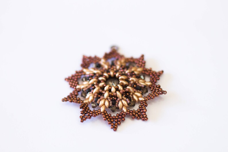 Op de afbeelding: Een bruine en gouden kralen sneeuwvlok ornament met een kristal in het midden.