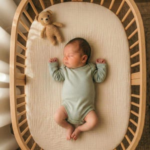 Puede incluir: Un bebé recién nacido con un body verde claro duerme plácidamente en una cuna ovalada de madera. Un pequeño osito de peluche de ganchillo descansa cerca del bebé. La cuna tiene un colchón de color claro y está cerca de una ventana.