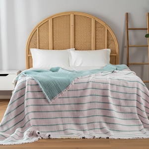 Könnte beinhalten: Ein Bett mit einem gewebten Rattan-Kopfteil, weißen Kissen und einer gestreiften Decke. Die Decke hat rosa und grüne Streifen und einen Fransenrand. Eine türkisfarbene Decke ist über das Bett drapiert. Eine Holzleiter und eine Topfpflanze befinden sich im Hintergrund.