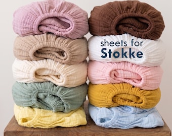 May include: A stack of ten folded muslin sheets in various colours including pink, brown, beige, cream, sage green, yellow, white, and light blue. The text "sheets for Stokke" is printed in dark blue on the white sheet in the right column.