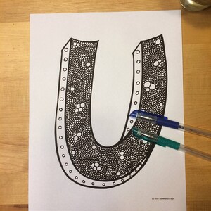 May include: A black and white printable colouring page featuring a large letter U with a detailed pattern of dots and circles. The letter U is surrounded by a white background.