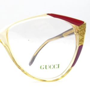 Puede incluir: Gafas Gucci vintage con montura amarillo claro, detalles en rojo y detalles dorados. Las lentes son transparentes y el logotipo de Gucci es visible. Las gafas tienen forma de ojo de gato.