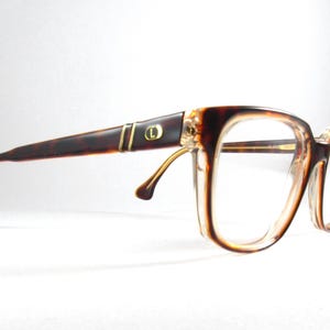 May include: Vintage eyeglasses with a brown tortoiseshell frame and clear lenses. The temples feature gold-colored accents and a small circular emblem. The glasses have a classic, rectangular shape.