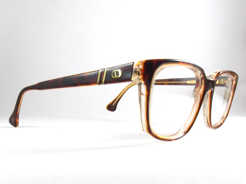 May include: Vintage eyeglasses with a brown tortoiseshell frame and clear lenses. The temples feature gold-colored accents and a small circular emblem. The glasses have a classic, rectangular shape.