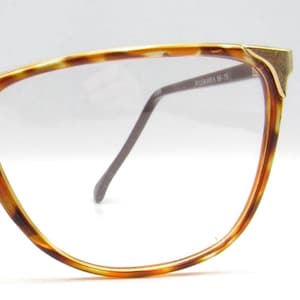 Op de afbeelding: Close-up van een bril met een montuur met schildpadpatroon. Het montuur heeft een gouden accent in de bovenhoek en donkergrijze veren. De glazen zijn helder. De tekst "NYLORARES 56-15" staat op de veer.