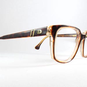 May include: Vintage eyeglasses with a tortoiseshell pattern on the arms and a clear, rectangular frame. The glasses have a gold accent near the hinges and a small gold emblem on the arm. The lenses are clear.