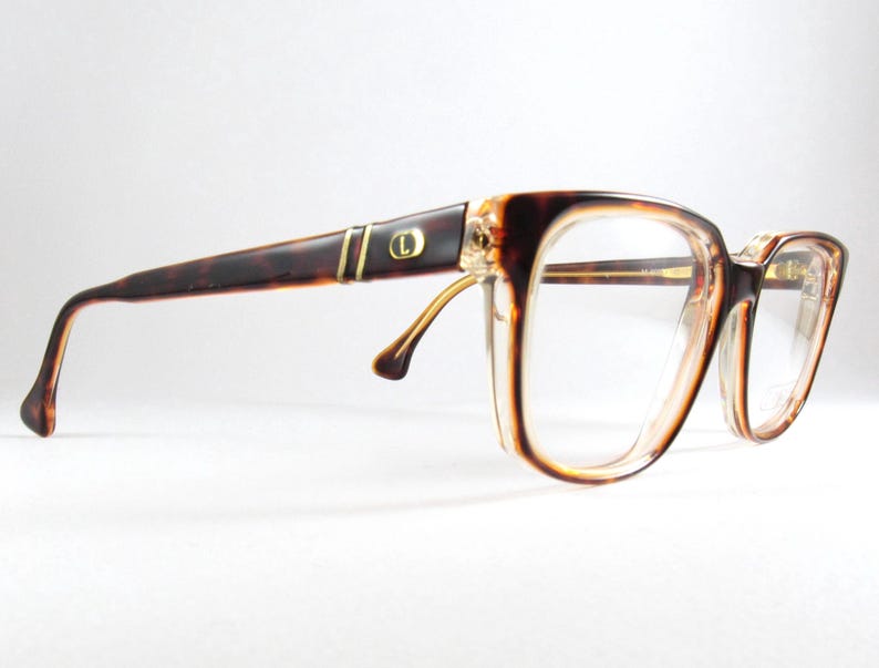 May include: Vintage eyeglasses with a tortoiseshell pattern on the arms and a clear, rectangular frame. The glasses have a gold accent near the hinges and a small gold emblem on the arm. The lenses are clear.