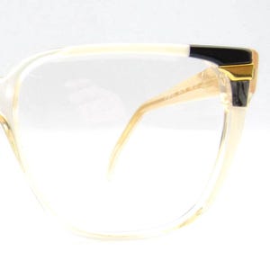 May include: Clear eyeglasses with a light beige frame. The temples are a light beige colour with a black accent and gold-coloured metal detailing. The lenses are clear, and the overall design is classic and elegant.